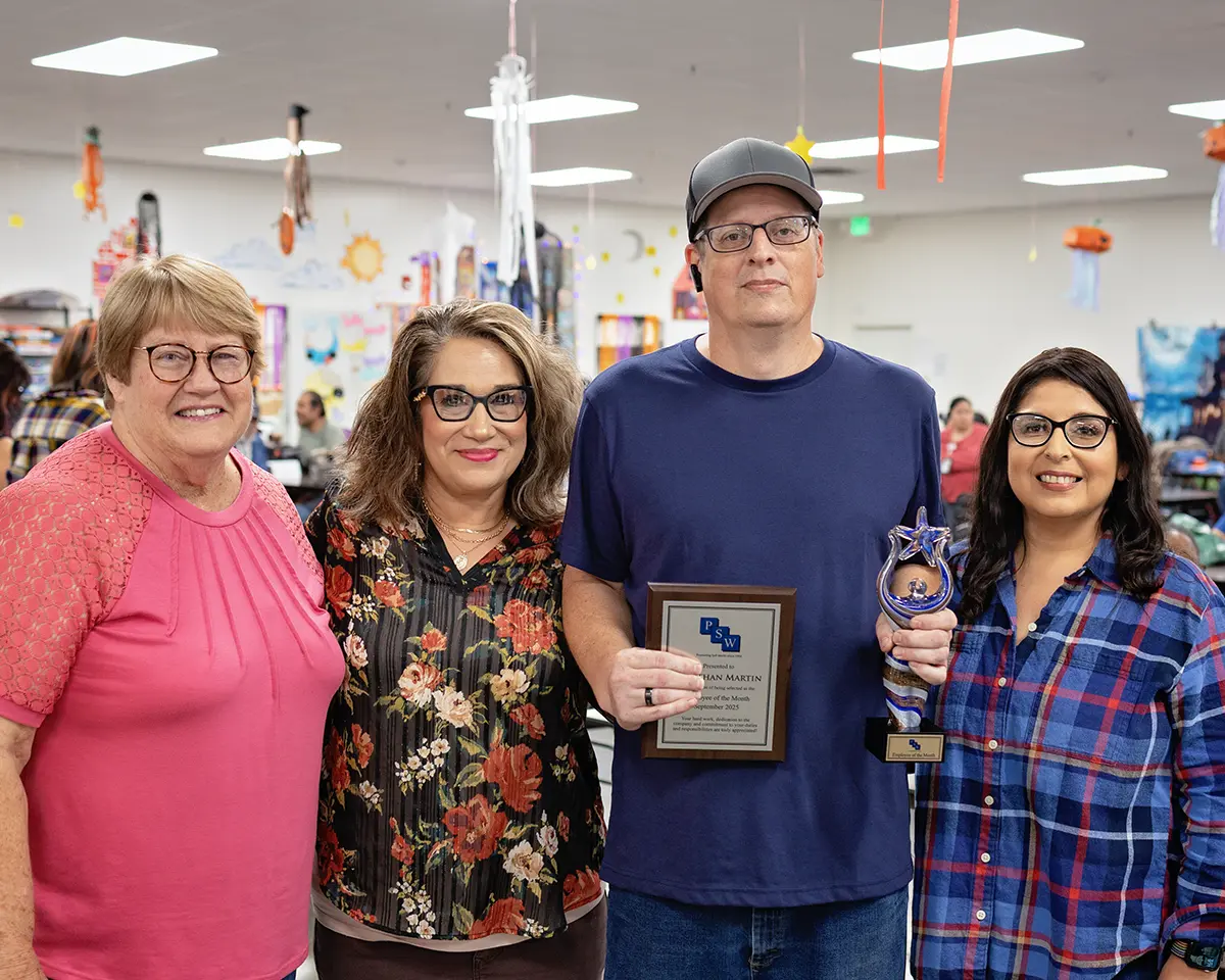 Four PSW staff members stand together smiling indoors, celebrating the September Employee of the Month. The award recipient holds a framed plaque and a blue star-shaped trophy, surrounded by supportive coworkers in a colorful, decorated room.